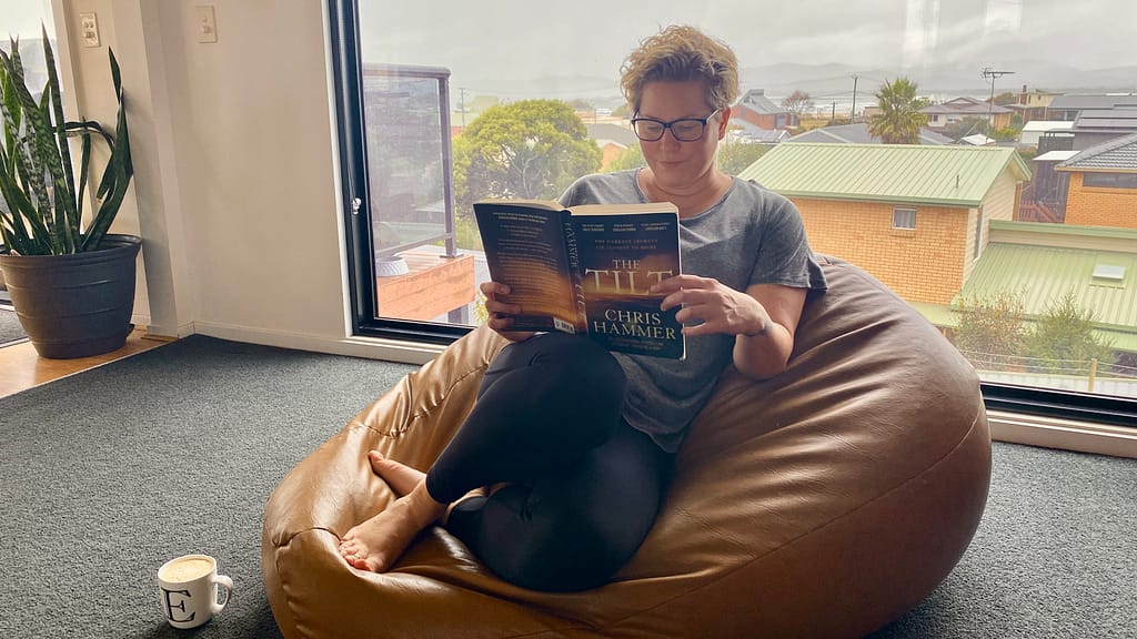 Em sits on a brown leather beanbag (made by her uncle) in her lounge room reading The Tilt by Chris Hammer. She has a white coffee mug with an E on it on the floor beside her. She is dressed for comfort, wearing tights, a grey top and bare feet. She also wears glasses. In the background is a large picture window looking out towards the water. It is a grey, overcast day.