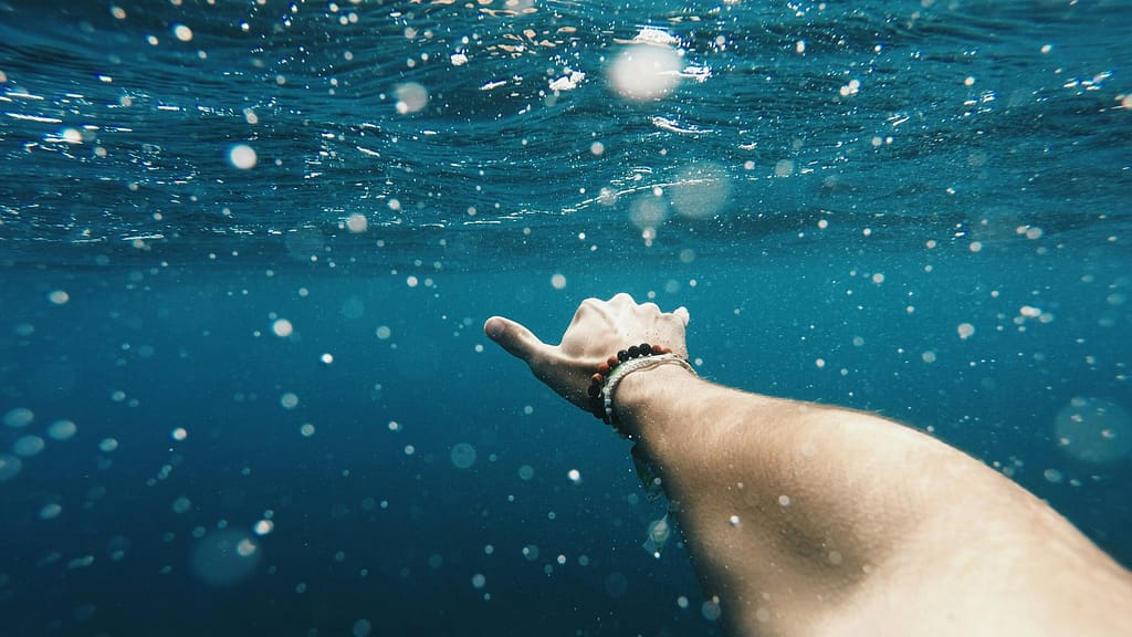 Underwater photo of the photographer's arm and hand. The water is blue and bubbly. They are wearing a beaded bracelet and doing the Hawaiian "Shaka".