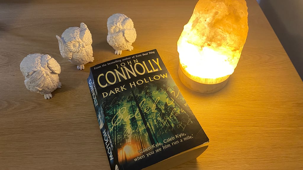 The book Dark Hollow by John Connolly sits on a desk beside a Himalayan salt lamp and three white ceramic owls that "see no evil, hear no evil, speak no evil".