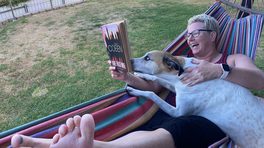 Em lies in a hammock trying to read Harlan Coben's One False Move. Luna, a white and tan greyhound, has decided this is a shared activity and is sniffing the book.
