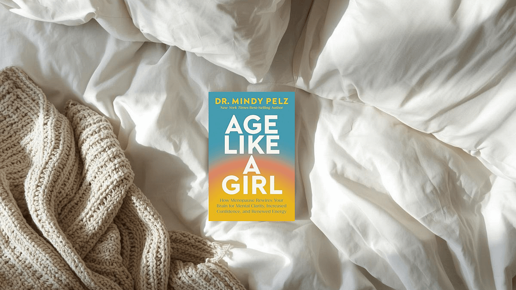Book cover of Age Like a Girl by Dr. Mindy Pelz resting on white bed sheets with a chunky beige knit blanket nearby.A comfortable spot for an Age Like a Girl review.