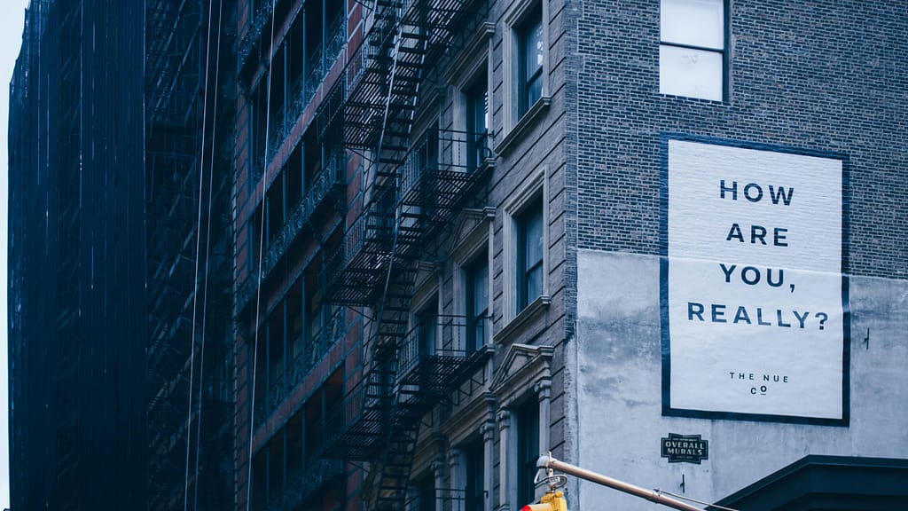 Urban building wall with a large white mural reading 'HOW ARE YOU, REALLY?' beside a fire escape in a blue-tinted city scene.