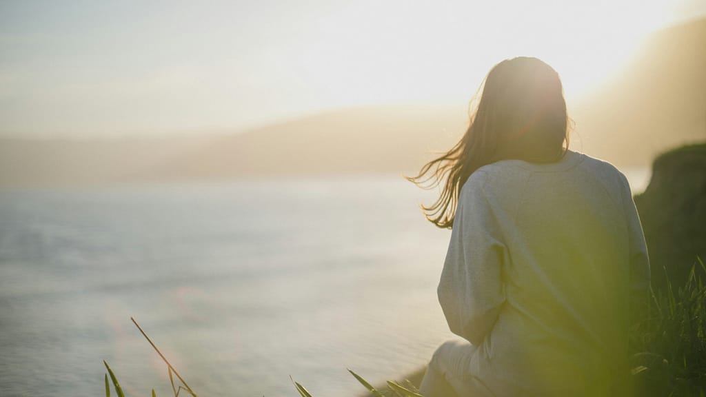 Back view of a person standing by the sea at sunset, hair blowing and wearing a light sweater.