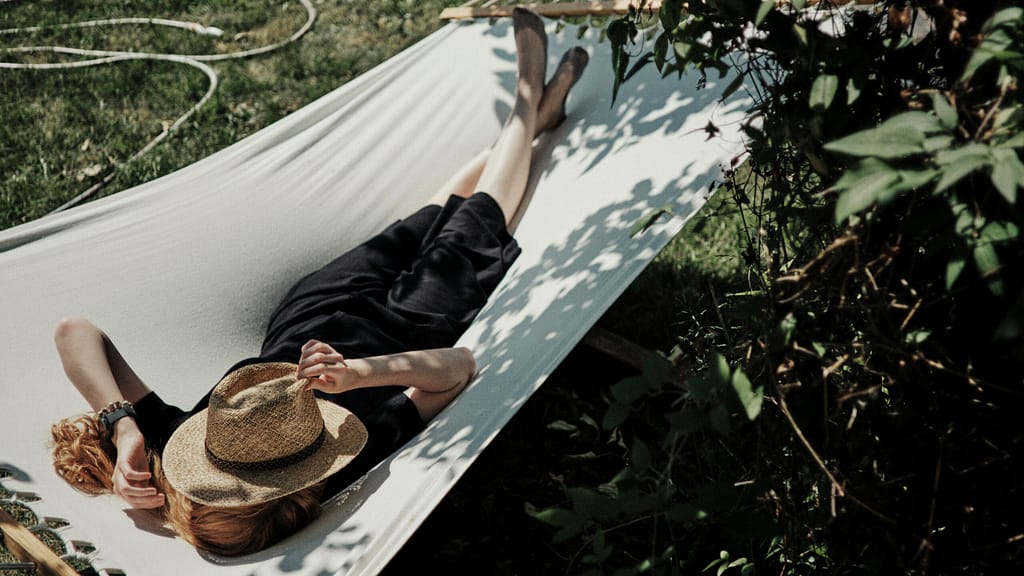 A woman lies in the sun in a hammock under a tree. She has a hat covering her face.