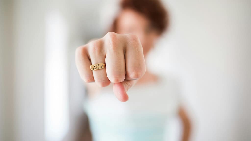 Close-up of a fist with a gold ring that reads 'I am bad ass' on the finger, blurred person in the background.
