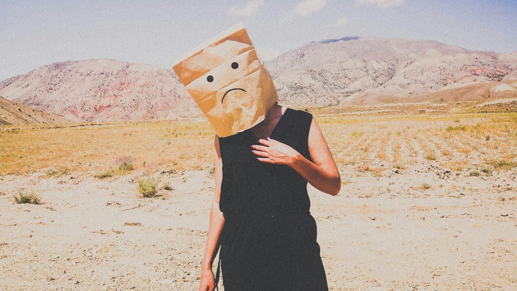 A woman stands in the desert with a brown paper bag on her head. The bag has a sad face drawn on it. Sometimes, people don't like you. But ignore them. They're d*cks.