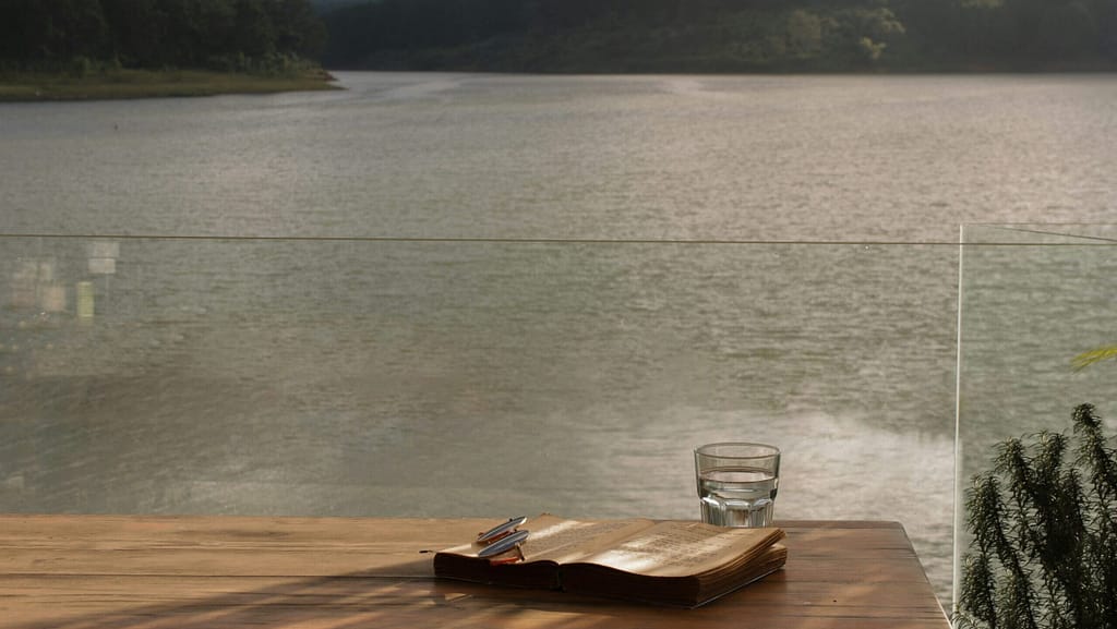A glass of water, a journal and some glasses sit on a timber table overlooking a serene lake.