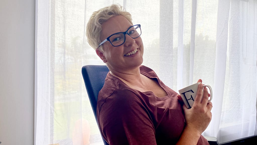 Life In Rough Draft writer Em sits on a chair, holding a mug with an E on it. She is side-on, but looking at the camera, leaning back in her chair. She has short blonde hair and blue glasses. She is smiling.