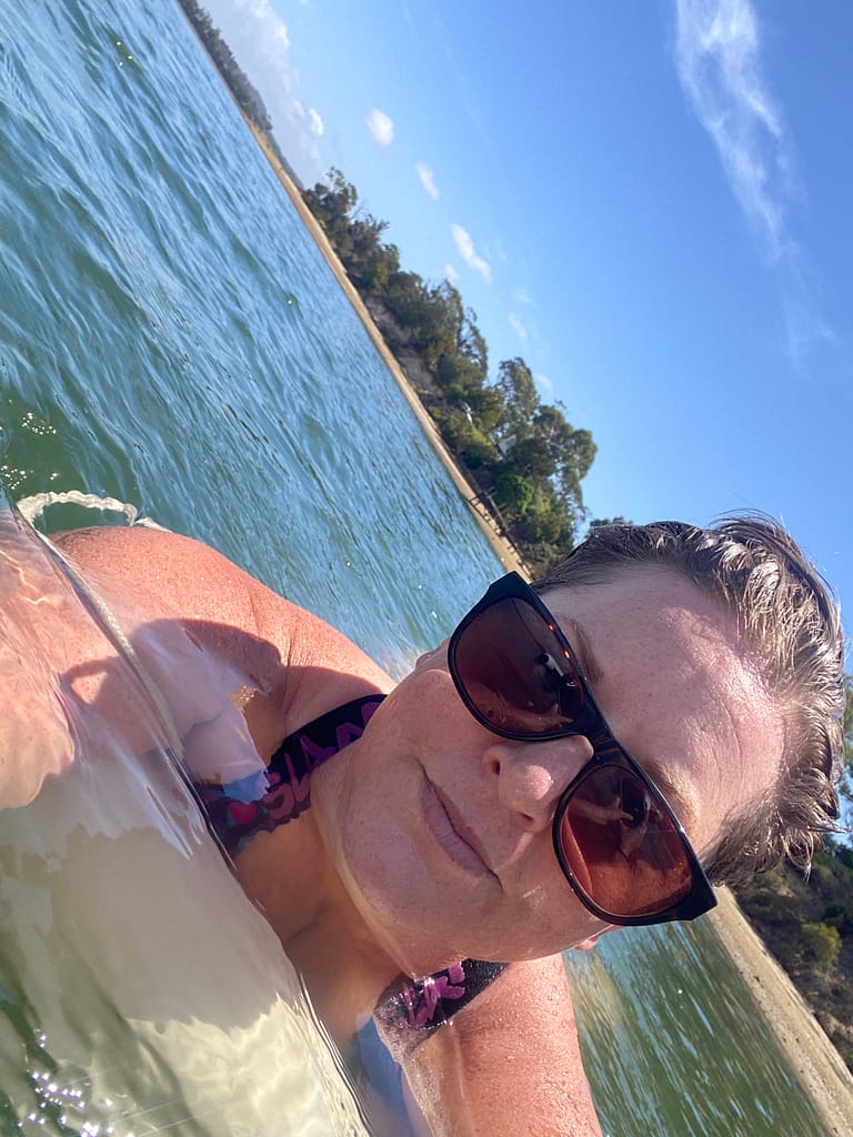Close up of Em floating in the water. She is wearing sunglasses and hanging off a float that is tied to the boat. The image is angled in a way that the background is crooked.