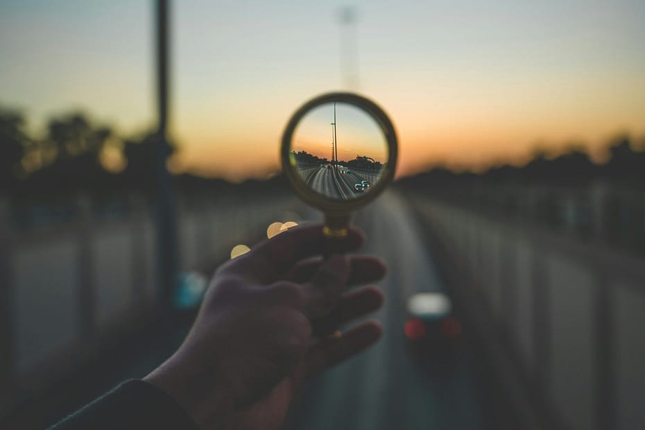 A hand holding a magnifying glass in front of a bridge at sunset. The centre of the magnifying glass is in focus, the rest of the image is blurry.