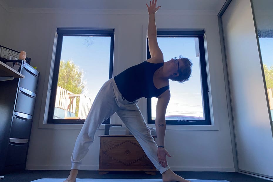Em on her yoga mat in triangle pose. Feet double-shoulder-width apart, arms open, upper body tilted to her left hand side. She is looking up towards her right hand which is vertical.