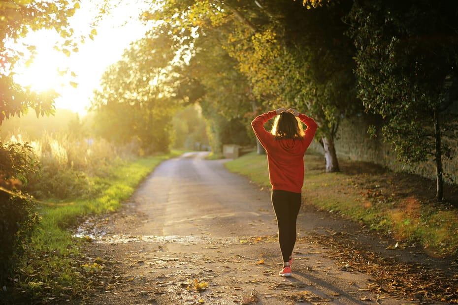 A woman walks along a country lane. Her hands are on her head and the sunrise is peeking through the trees.