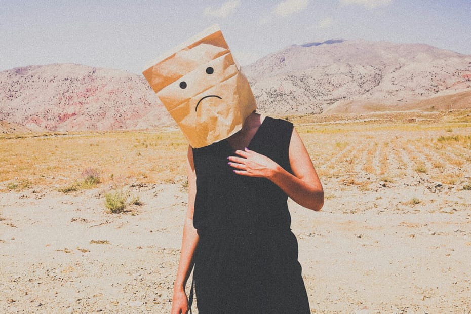 A woman stands in the desert with a brown paper bag on her head. The bag has a sad face drawn on it.