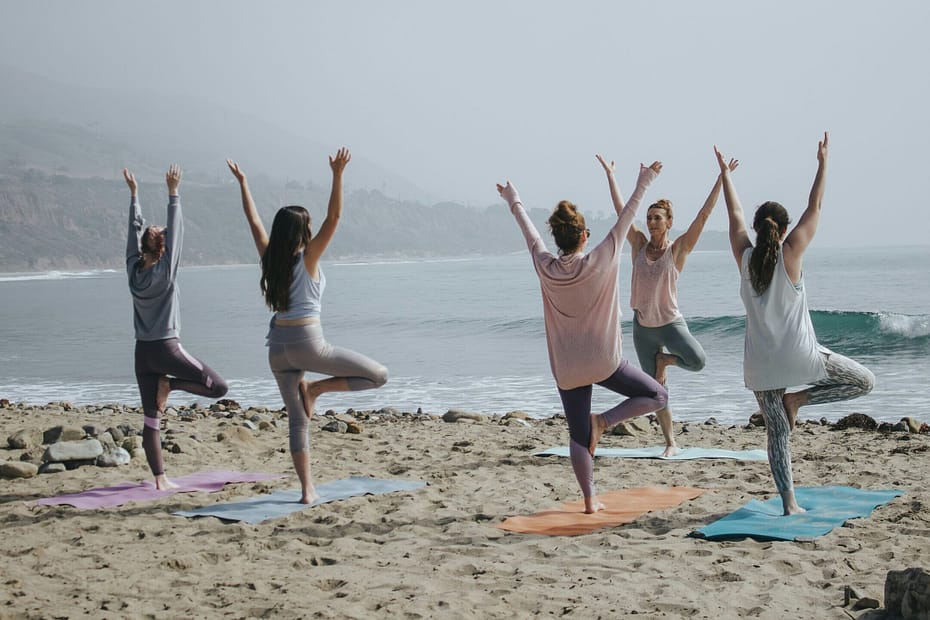 A group of five women do yoga on a beach. It's a misty day and there are waves in the background.