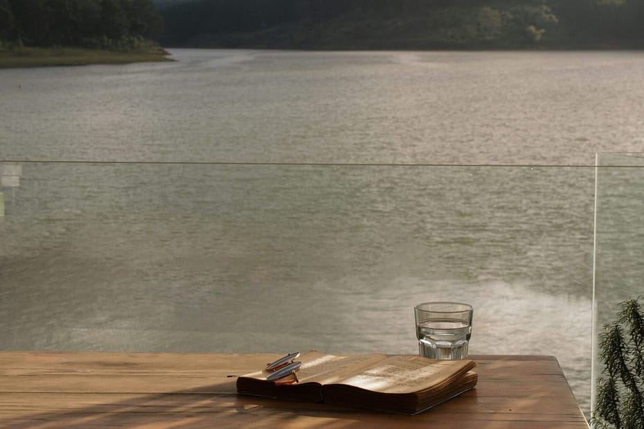 A glass of water, a journal and some glasses sit on a timber table overlooking a serene lake.