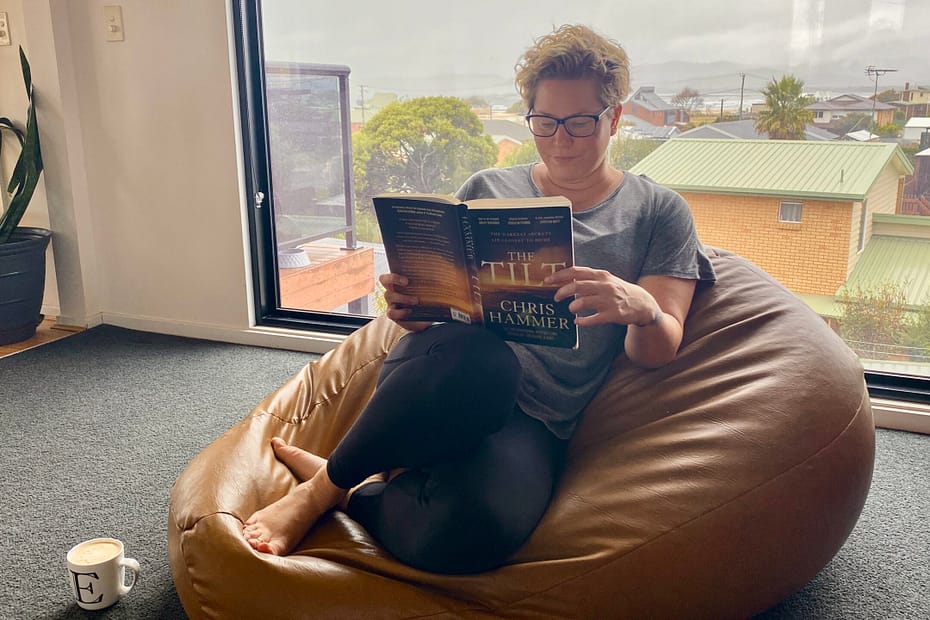 Em sits on a brown leather beanbag (made by her uncle) in her lounge room reading The Tilt by Chris Hammer. She has a white coffee mug with an E on it on the floor beside her. She is dressed for comfort, wearing tights, a grey top and bare feet. She also wears glasses. In the background is a large picture window looking out towards the water. It is a grey, overcast day.