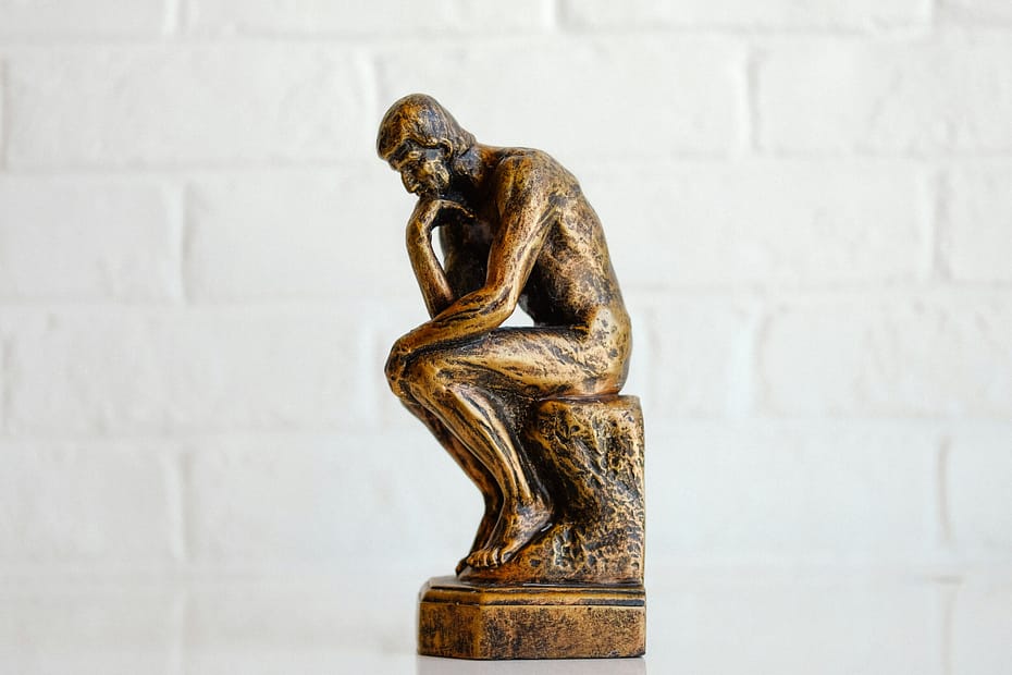 A tiny replica statue of The Thinker sits on a white table in front of a white brick wall. Maybe he's battling his own negative self-talk.