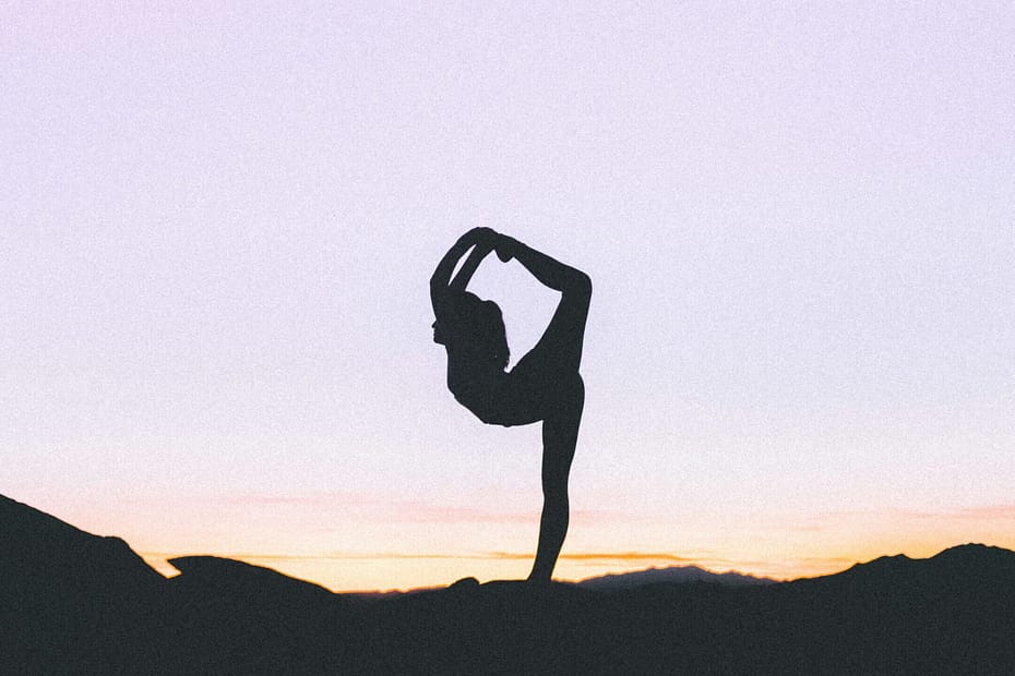 woman silhouetted against a purple and orange sunset stands on one leg with her other leg bent up behind her and over her head. This woman is clearly not Em.