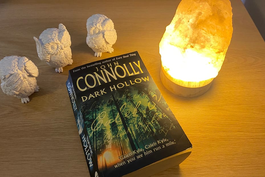 The book Dark Hollow by John Connolly sits on a desk beside a Himalayan salt lamp and three white ceramic owls that "see no evil, hear no evil, speak no evil".