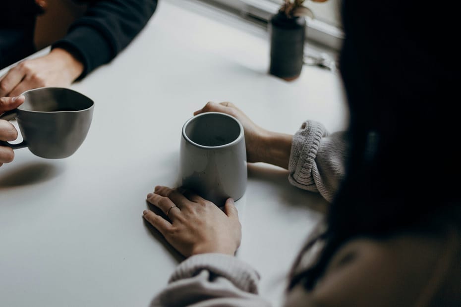 Two friends sit having coffee. We can only see their hands and coffee cups, but we can tell they are showing up for each other.