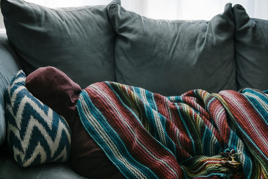 A person lays on a blue-green couch under a colourful blanket. They have their back to us.