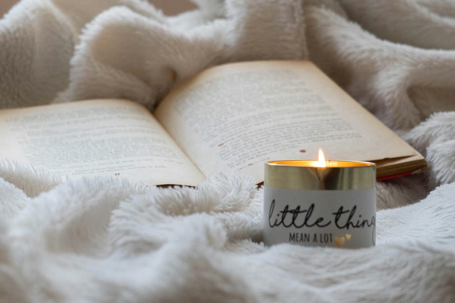 A white candle sits burning on a cosy blanket, next to a book.