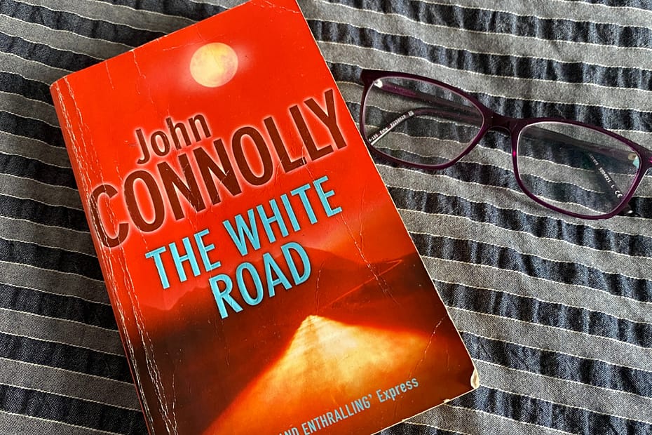 The White Road by John Connolly and a pair of glasses on a bed.