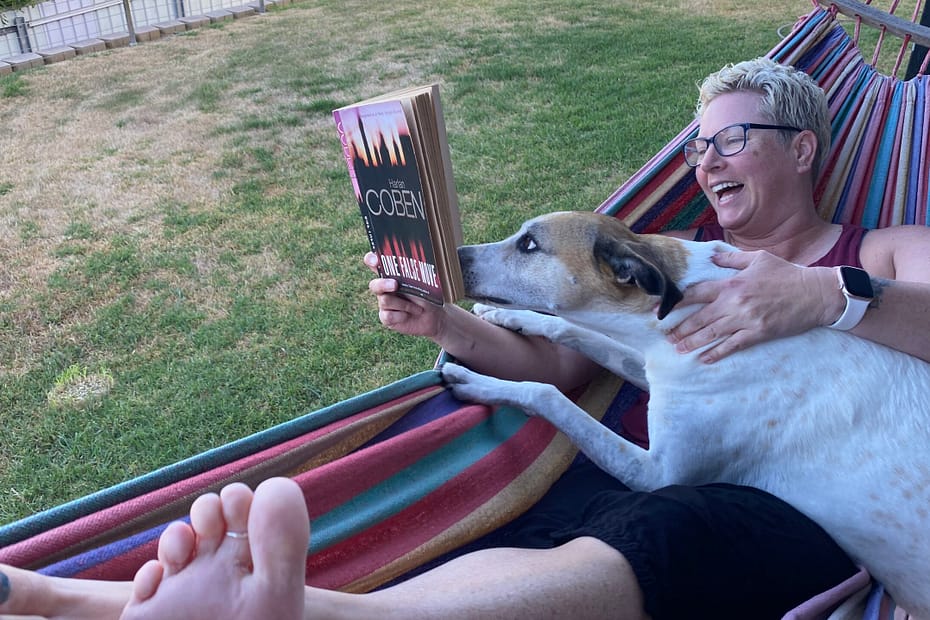 Em lays in a hammock reading Harlan Coben's One False Move. Luna, a white and tan greyhound, has jumped into her lap and is nosing at the book as if she's reading it intently. Em is laughing.