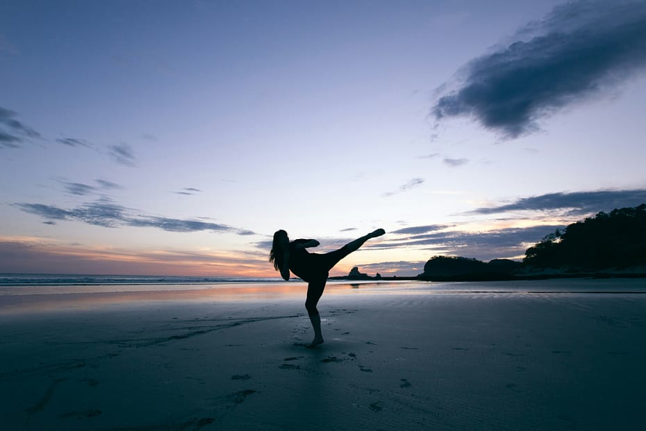 A woman practices a martial arts side kick on the beach at sunset.