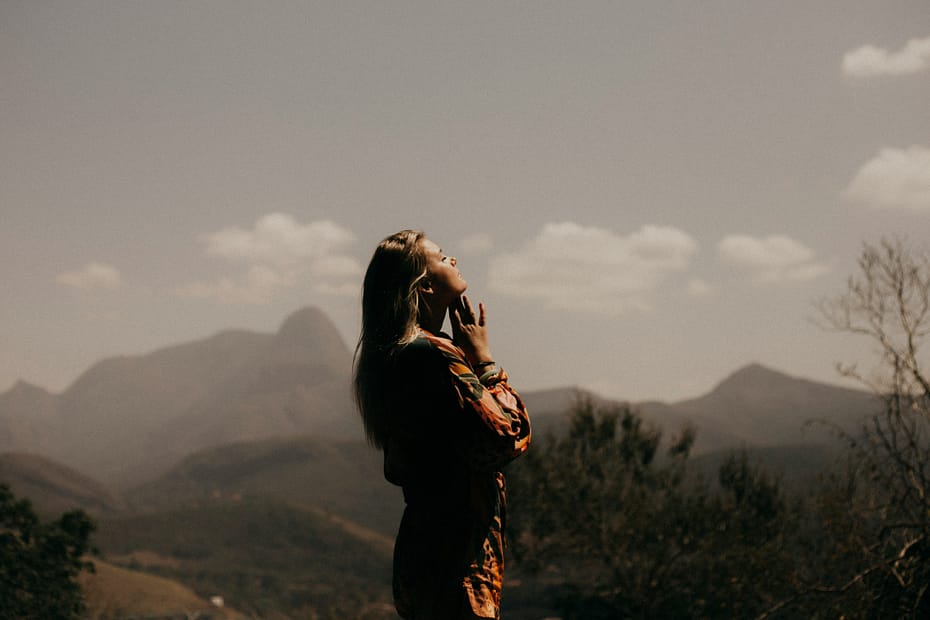 A woman stands in nature, a mountain range behind her, with her face to the sky and her hands together as if in prayer.