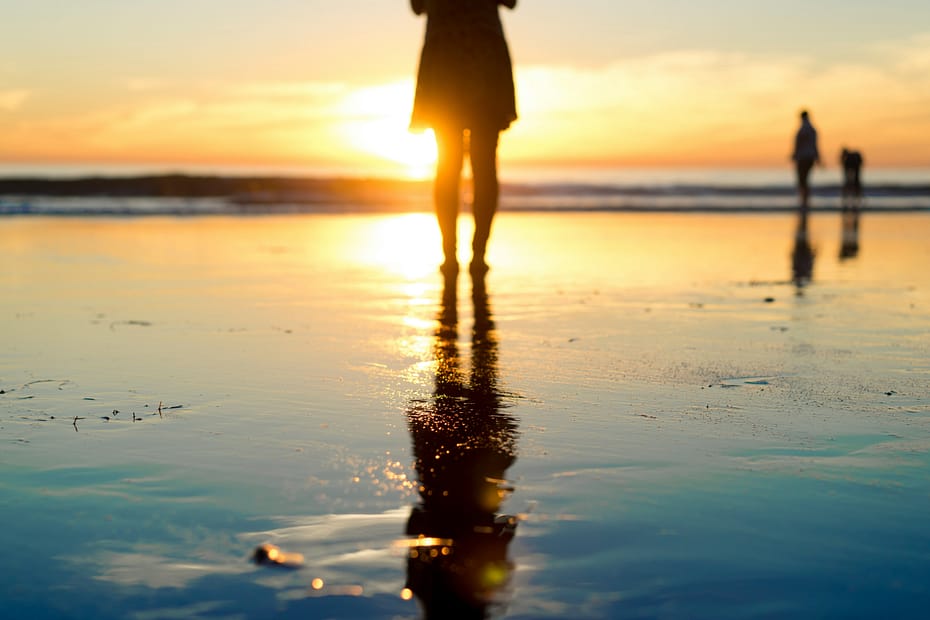 A woman stands on a beach in front of a sunset. It is a low shot, showing her back and legs and the sunset is reflecting off the wet sand.
