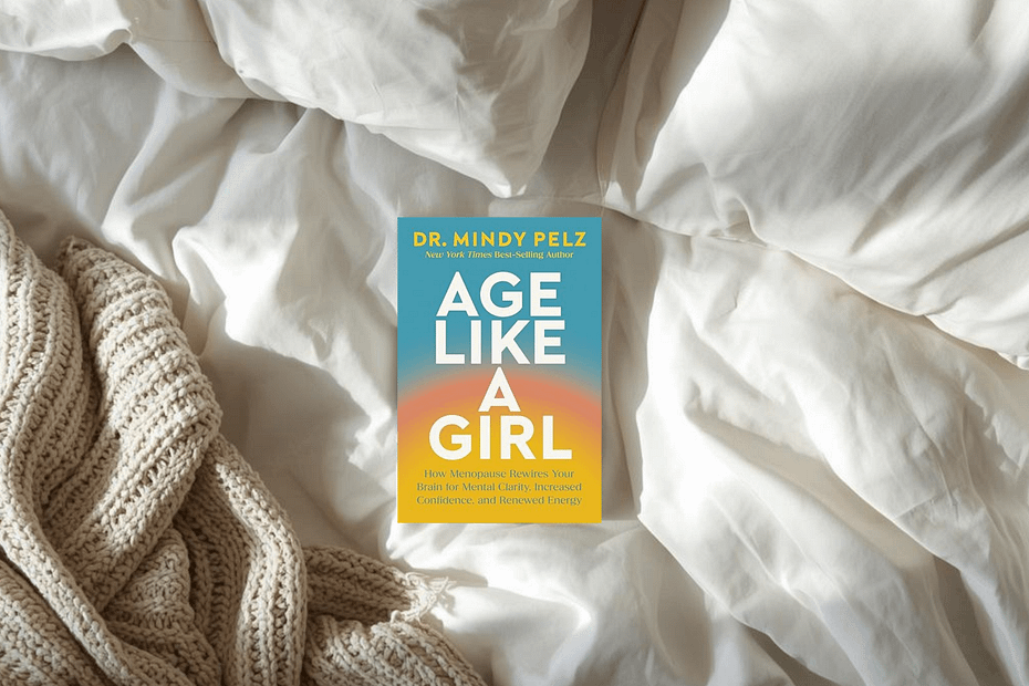 Book cover of 'Age Like a Girl' by Dr. Mindy Pelz resting on white bed sheets with a chunky beige knit blanket nearby.