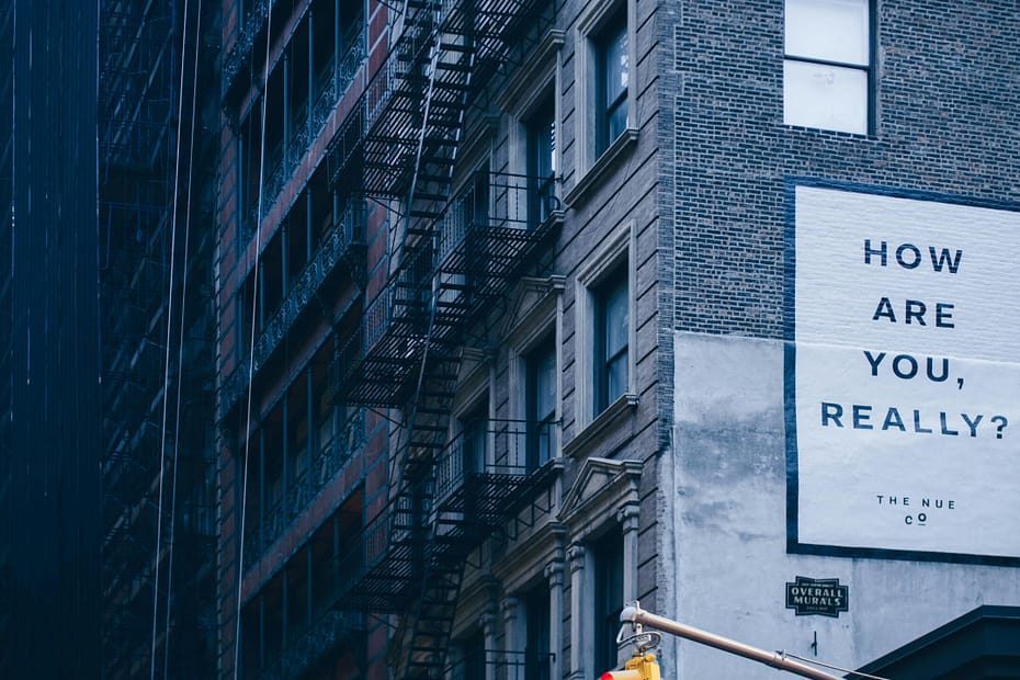 Urban building wall with a large white mural reading 'HOW ARE YOU, REALLY?' beside a fire escape in a blue-tinted city scene.