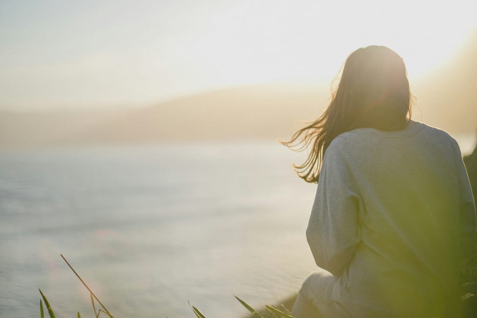 Back view of a person standing by the sea at sunset, hair blowing and wearing a light sweater.