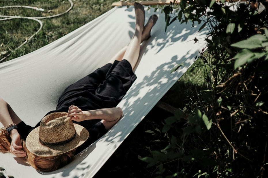 A woman lies in the sun in a hammock under a tree. She has a hat covering her face.