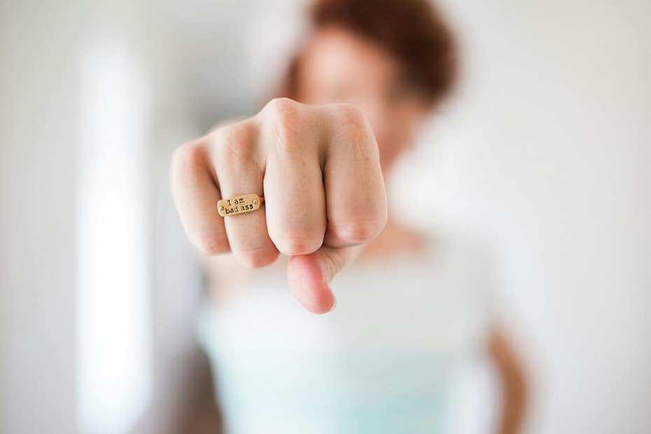 Close-up of a fist with a gold ring that reads 'I am bad ass' on the finger, blurred person in the background.
